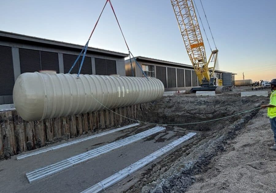 Crawler crane lowering a fiberglass fuel storage tank into an excavation at dusk